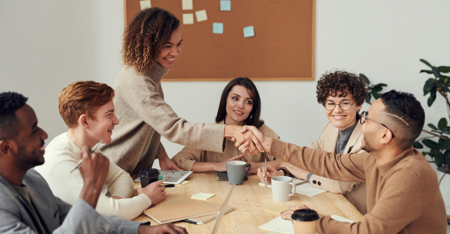 A diverse group of professionals engaging in a collaborative meeting, with a woman in a beige sweater shaking hands with a colleague, symbolizing workplace alignment, ethical leadership, and inclusive teamwork. Office setting with coffee cups, notebooks, and a corkboard with notes in the background, reflecting a warm and engaging business culture.