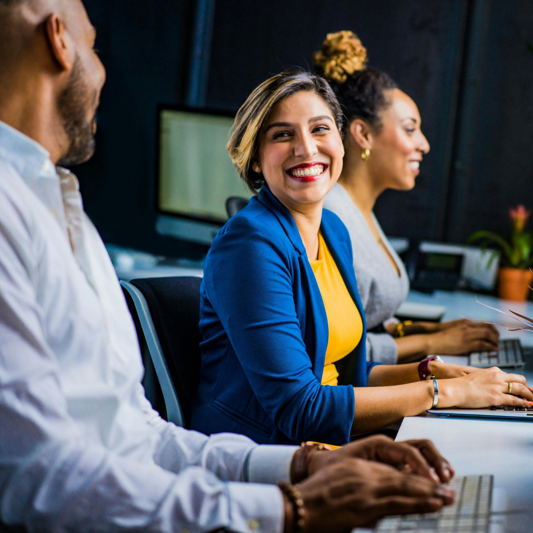 A diverse group of professionals working in a modern office, with a smiling woman in a blue blazer engaging in conversation. The scene reflects collaboration, positive workplace culture, and professional growth in an inclusive environment.